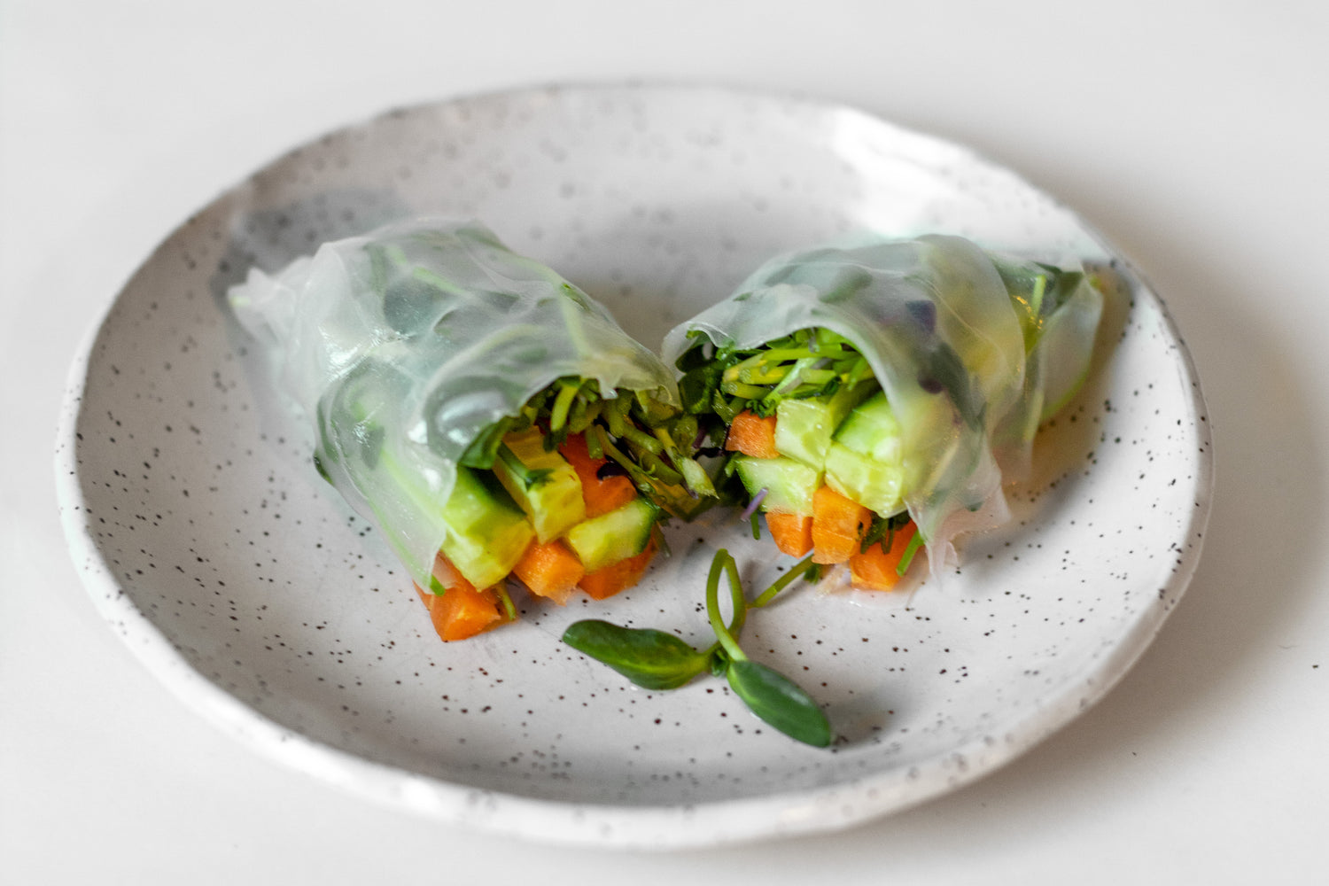 Image of a rice paper roll on a plate against a white background. The roll is cut in half and filled with an assortment of vegetables, including collar greens, cucumber, carrots, and microgreens.