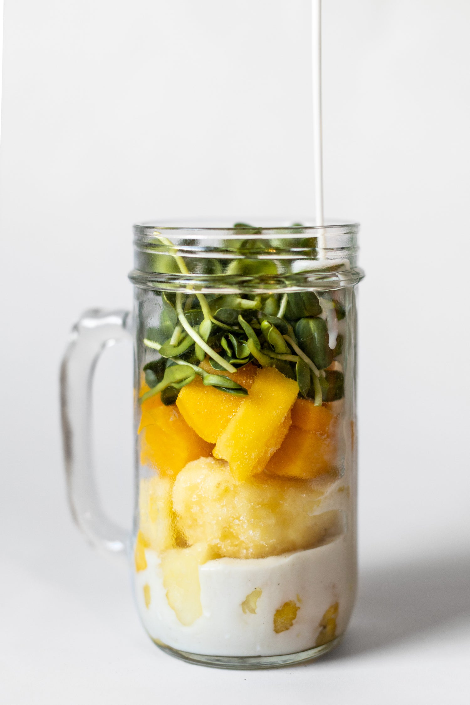 Image of smoothie cup against a white background. Milk, bananas, mango, and microgreens are layered in the cup.
